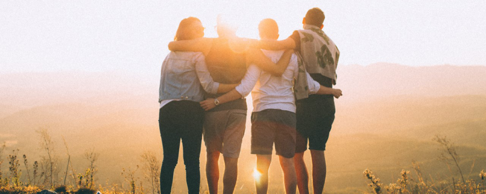 Four young adults stand together with arms around each other, watching the sunset over a scenic mountain landscape.