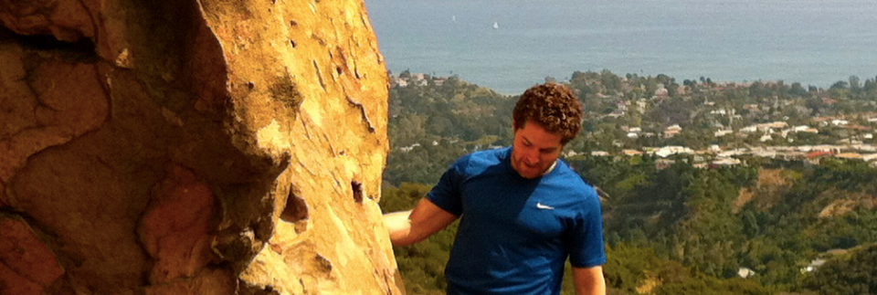 Man rock climbing outdoors near Malibu with ocean and coastal homes in the background on a sunny day.