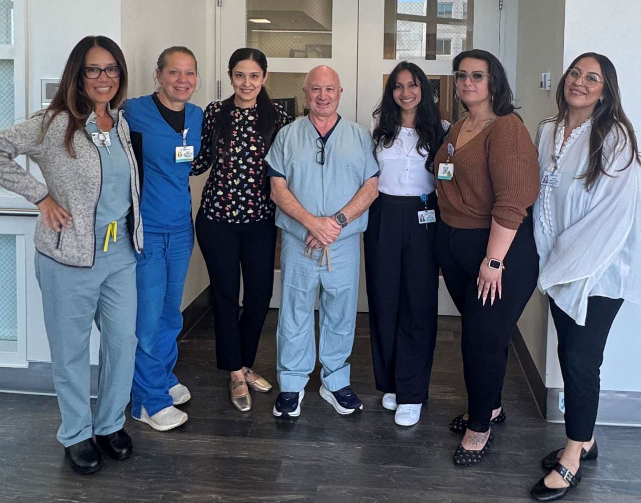 Group of seven healthcare professionals smiling together indoors, with some in scrubs and others in business casual attire.
