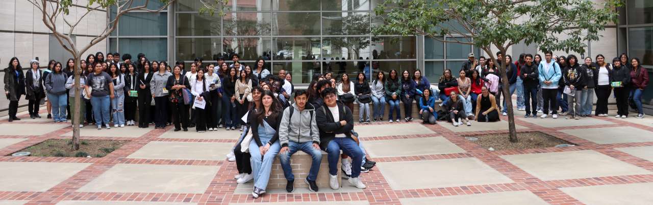 A large group of diverse students and faculty pose for a photo outside a building with large windows. A few people are sitting in the foreground.