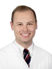 A headshot of Adrien Holzgreve, a man with short brown hair, smiling and wearing a white lab coat and a striped tie.