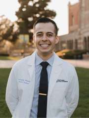 Person in a white coat and tie standing on a campus lawn at sunset.