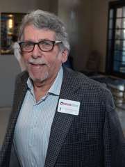Gary Edelstone in a checkered blazer wearing a name badge, walking in a well-lit indoor space.