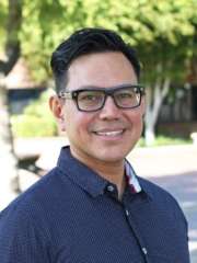 A man stands outdoors in a casual shirt, surrounded by greenery.