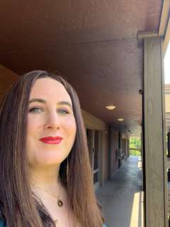 Chloe smiling with long brown hair and red lipstick looking at the camera on a porch with pillars.