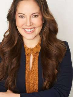 Person with long wavy brown hair wearing a navy blazer over a mustard lace blouse.