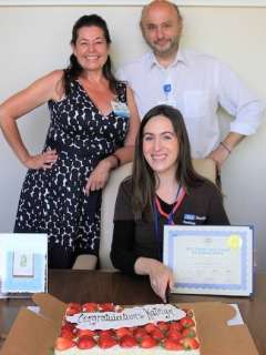 Three people at a table with a congratulatory cake, a certificate, and greeting cards displayed.