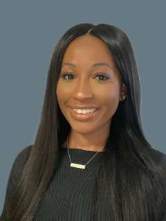 Brittany Tarrant with long straight black hair, wearing a black top and necklace, smiles against a plain gray background.