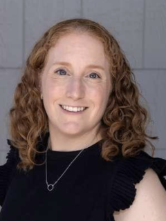 A woman with red curly hair, wearing a black skirt with a necklace, smiling in front of a tiled background