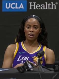 A headshot of WNBA player Jordin Canada in a purple and gold Lakers jersey, speaking at a press conference with a UCLA Health banner in the background