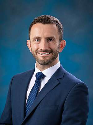 Smiling man in navy suit and tie against blue studio background.