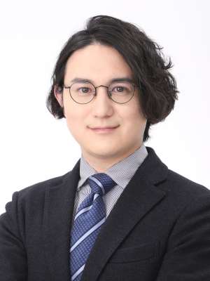 A headshot of Koichiro Kimura with dark, wavy hair, round glasses, and smiling, wearing a dark suit, light shirt, and striped tie.
