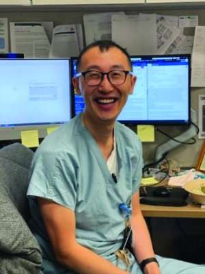 Smiling person in teal medical scrubs seated at a desk with monitors