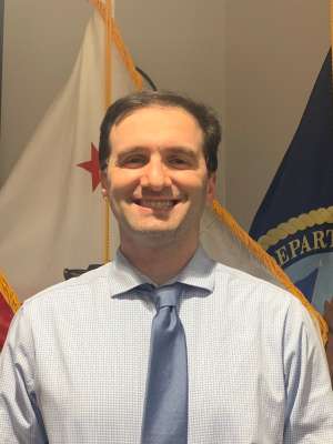 Smiling man in shirt and tie with U.S. flag and Veterans Affairs flag behind him.