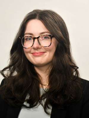 Smiling woman with long dark hair, glasses, and black blazer on neutral background