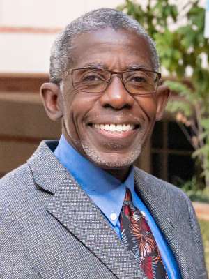 Headshot of Doctor Dale Abel in a grey suit, wearing glasses and standing in front of a brick building with trees to the right.