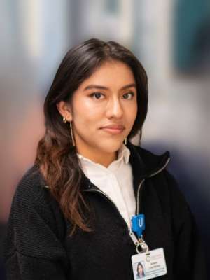  A professional photo of Maria Contreras, a woman with long dark hair, wearing a black zippered top over a white shirt, and an ID badge.