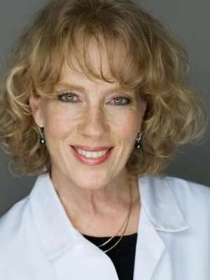 Person with curly blond hair wearing white lab coat and gold necklace