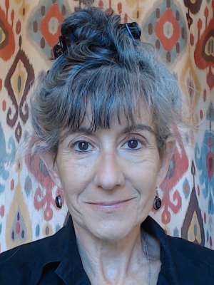 A person with gray hair tied up, wearing a black shirt, in front of a colorful patterned background.