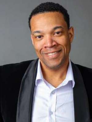 Smiling man in black blazer and white shirt against gray background.