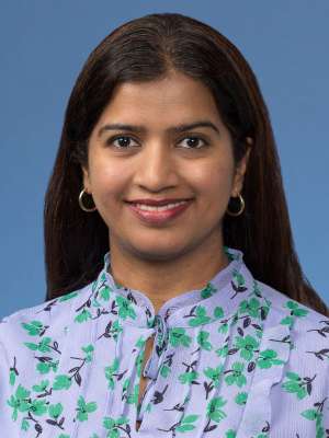 Headshot of a woman with long dark hair, wearing a floral blouse against a blue background.
