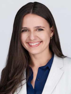 Smiling woman with long dark hair in white blazer and navy blouse, headshot.