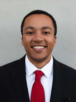 Smiling man in black suit, white shirt and red tie against gray background.