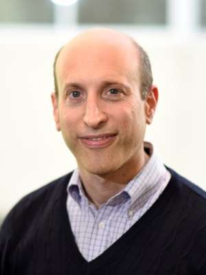 Man in a dark sweater stands in front of a bright, blurred background.