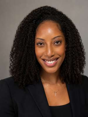 Smiling woman with curly hair in black blazer headshot on gray background.