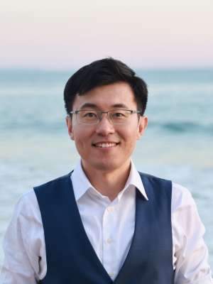 Smiling person wearing glasses, white shirt and navy vest at the beach.