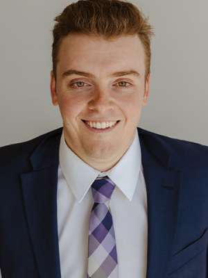 Smiling man in navy suit and purple plaid tie on neutral background.