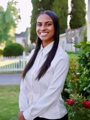 A professional headshot of Lela Theus, a smiling woman with long, straight dark hair, wearing a white long-sleeved shirt outdoors.