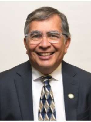 A man with short grey hair, wearing a suit and tie with glasses, smiling in front of a blank background