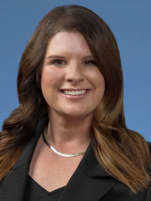 Emily Stimpson with long wavy hair, wearing a black blazer and silver necklace, posed confidently against a blue background.
