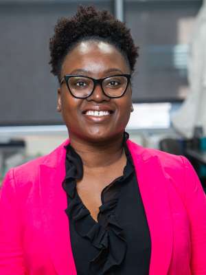 Rhonda Souvenir wearing black sunglasses, black ruffle blouse, and pink blazer. Background is blurry.