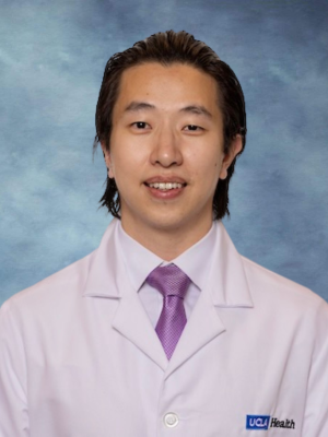 A headshot of Dr. Jerry Lee who has dark brown hair and is wearing a white lab coat, purple tie and smiling
