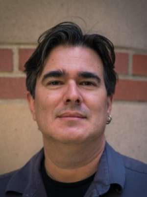 Head-and-shoulders portrait of a man with dark hair and earring against a brick wall