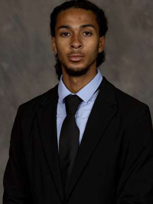 Headshot of a man wearing a black suit, light blue shirt, and black tie against gray backdrop.