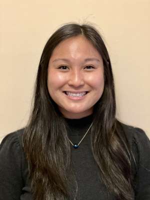 Smiling woman with long dark hair wearing black top and necklace on beige background.