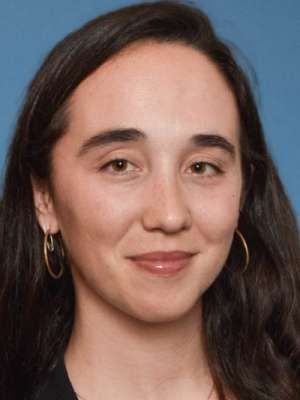 Headshot of a woman with dark hair, hoop earrings, neutral smile on blue background.