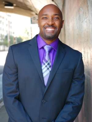 Chayil Champion headshot wearing suit, purple shirt and plaid tie, smiling
