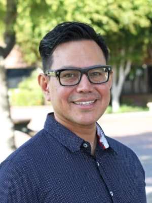 A man stands outdoors in a casual shirt, surrounded by greenery.