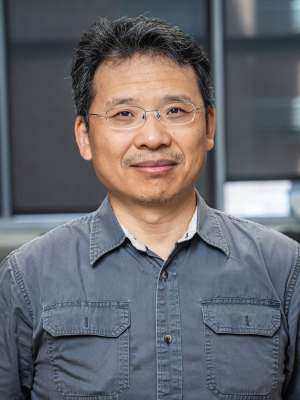 Headshot of Doctor Zhang wearing glasses and a grey button-down shirt, the background is blurry