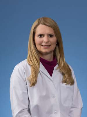 Blonde woman, Kailee Gaffny, wearing a white lab coat over a maroon top, stands in a professional, confident pose against a blue background.