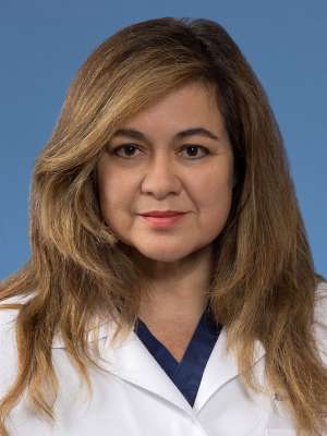 Professional headshot Ana Valenzuela with long, brown, layered hair, wearing a white medical coat over blue scrubs with a calm, confident expression.
