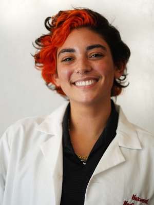 Smiling person with short red and black hair wearing a white lab coat
