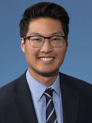 Professional headshot of person in suit and tie wearing glasses, smiling on blue background