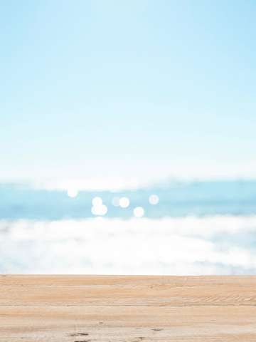 A blurry image of an ocean on a sunny day with a light-colored wooden table in the foreground.