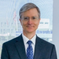 A man with short grey hair, wearing glasses with a suit and tie, posing in front of a building window