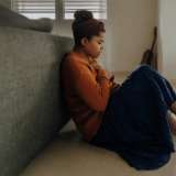 A depressed younger woman sits against her bed.